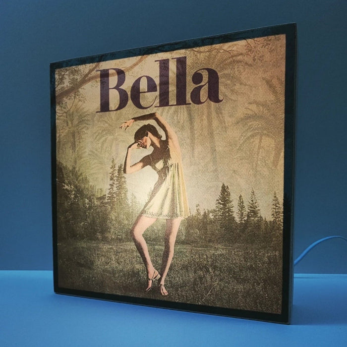 Caja de luz de madera y papel con la imagen de una bailarina frente a un bosque. Mientras está apagada sólo aparece la bailarina y el fondo vegetal, cuando está encendida aparece en la parte superior de la imagen la palabra "bella". Caja de luz hecha de forma manual.