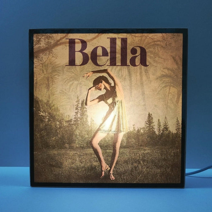 Caja de luz de madera y papel con la imagen de una bailarina frente a un bosque. Mientras está apagada sólo aparece la bailarina y el fondo vegetal, cuando está encendida aparece en la parte superior de la imagen la palabra "bella". Caja de luz hecha de forma manual.