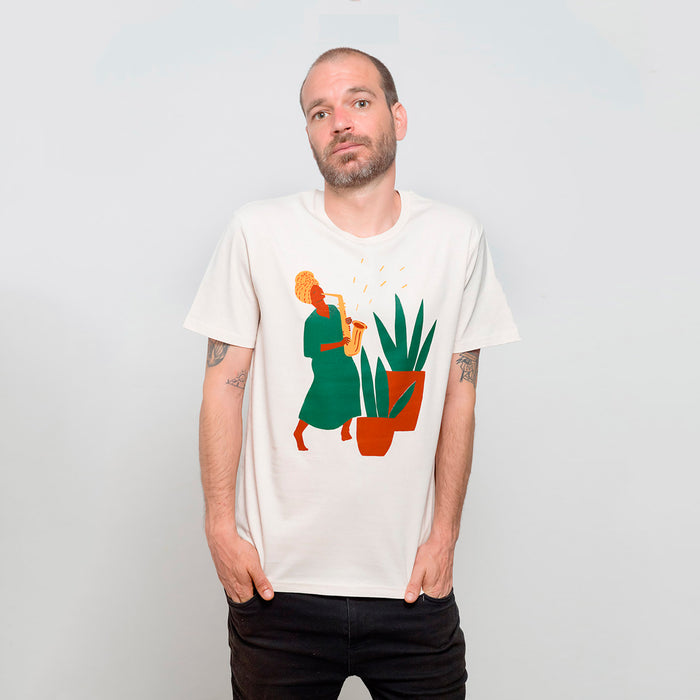 Camiseta blanca con ilustración de mujer negra tocando el saxofón junto a unas macetas grandes con plantas en tonos tierra, verdes y amarillos.