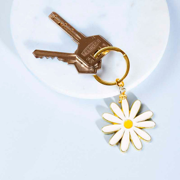 Llavero de metal dorado con charm en forma de margarita con dos llaves sobre un plato blanco y  fondo azul claro.