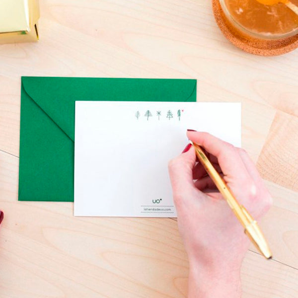 Fotografía de mano escribiendo con un boligrafo dorado en el reverso de una postal en color blanco con dibujos de árboles geométricos en verde sobre un sobre de color verde en una mesa de madera clara.