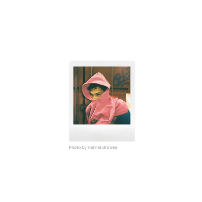 Fotografia polaroid go de un hombre con sudadera rosa con capucha ante una puerta metalica tomada por Harriet Browse