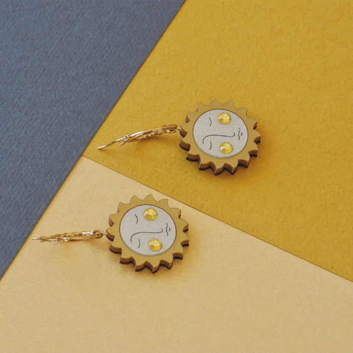 Pendientes de aro con charm de metacrilato en forma de sol con rostro y cristal de swarovski en las mejillas en plano lateral en fondo de tonos amarillos y azules