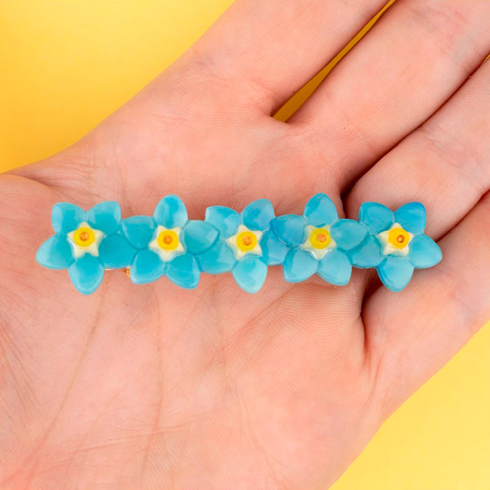 Pasador de pelo en forma de fila de flores nomeolvides azules con detalles en blanco y amarillo. Fabricado en acetato de celulosa reciclado. Vista sobre mano.