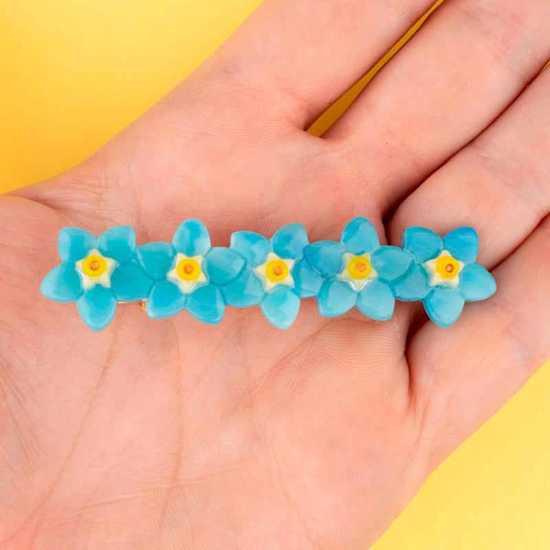 Pasador de pelo en forma de fila de flores nomeolvides azules con detalles en blanco y amarillo. Fabricado en acetato de celulosa reciclado. Vista sobre mano.