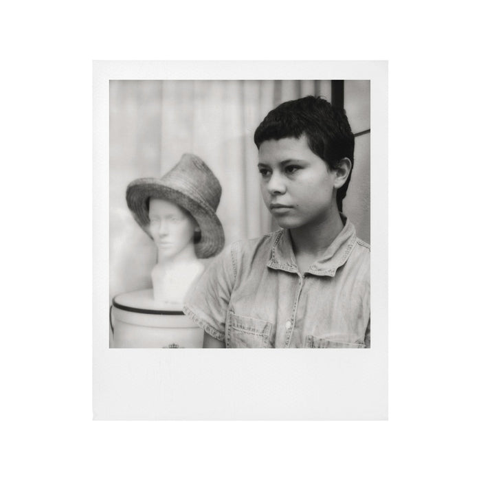 Fotografía polaroid i-Type en blanco negro de una mujer racializada con pelo corto junto a un busto con sombrero