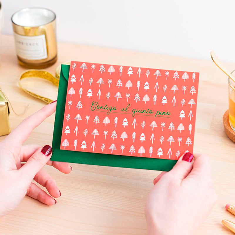 Fotografía de manos de mujer con uñas pintadas de rojo sujetando una postal de color rojo con estampado de árboles geométricos y el texto "contigo al quinto pino" y sobre de color verde.