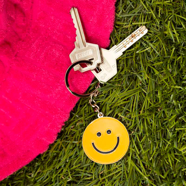 Llavero plateado de metal con charm de carita sonriente redonda amarilla con dos llaves sobre hierba.