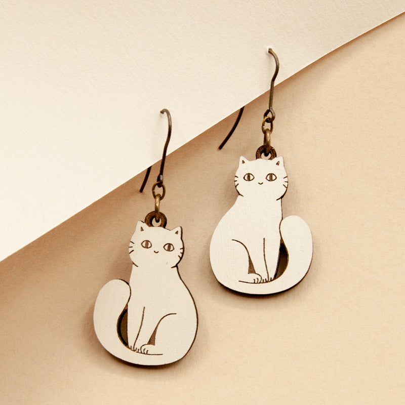 Pendientes de gancho con charm de gato blanco sentado en una cartulina de color beige sobre fondo beige.