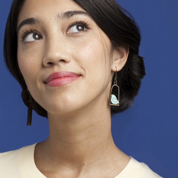 Modelo con Pendientes desparejados en forma de pájaro azul en un columpio y jaula de pájaro en fondo azul.
