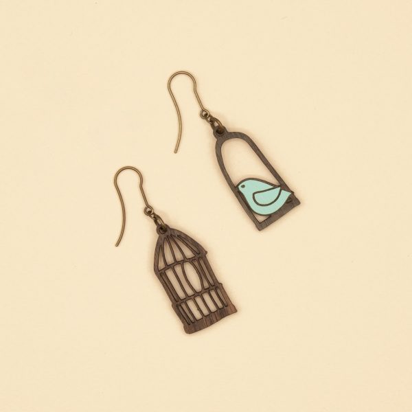 Pendientes desparejados en forma de pájaro azul en un columpio y jaula de pájaro sobre fondo crema.