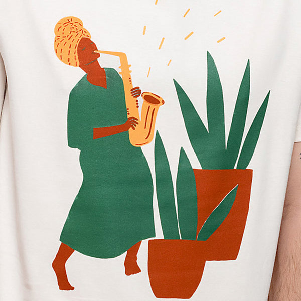 Camiseta blanca con ilustración de mujer negra tocando el saxofón junto a unas macetas grandes con plantas en tonos tierra, verdes y amarillos. 