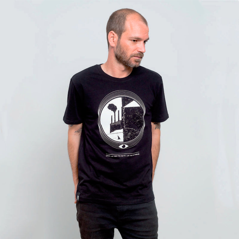 Camiseta negra con ilustración en circular de una fábrica con chimenea junto al a playa y un faro iluminando el cielo nocturno junto con el texto "When we heal the earth we heal ourselves", todo en tonos blancos.