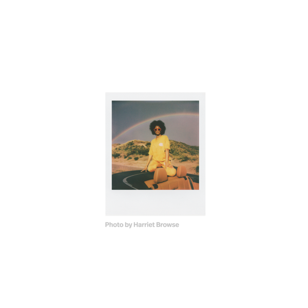 Fotografia polaroid go de una mujer negra vestida de amarillo con gafas de sol rojas sobre la parte trasera de un descapotable en un fondo de naturaleza con arcoiris tomada por Harriet Browse