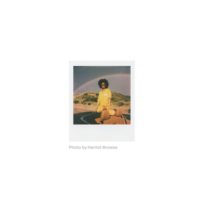 Fotografia polaroid go de una mujer negra vestida de amarillo con gafas de sol rojas sobre la parte trasera de un descapotable en un fondo de naturaleza con arcoiris tomada por Harriet Browse