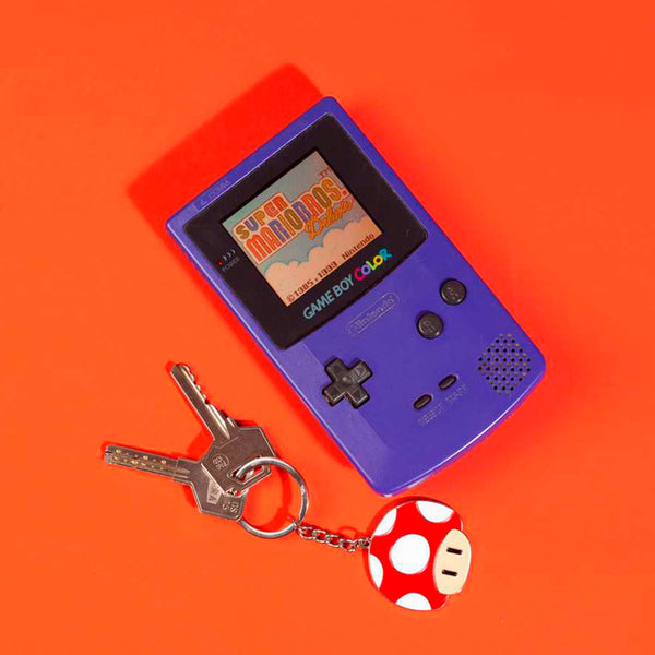 Llavero metálico plateado en forma de seta 1up de Super Mario con dos llaves junto con Game Bo Color color azul con videojuego Super Mario Bros Deluxe en fondo rojo.