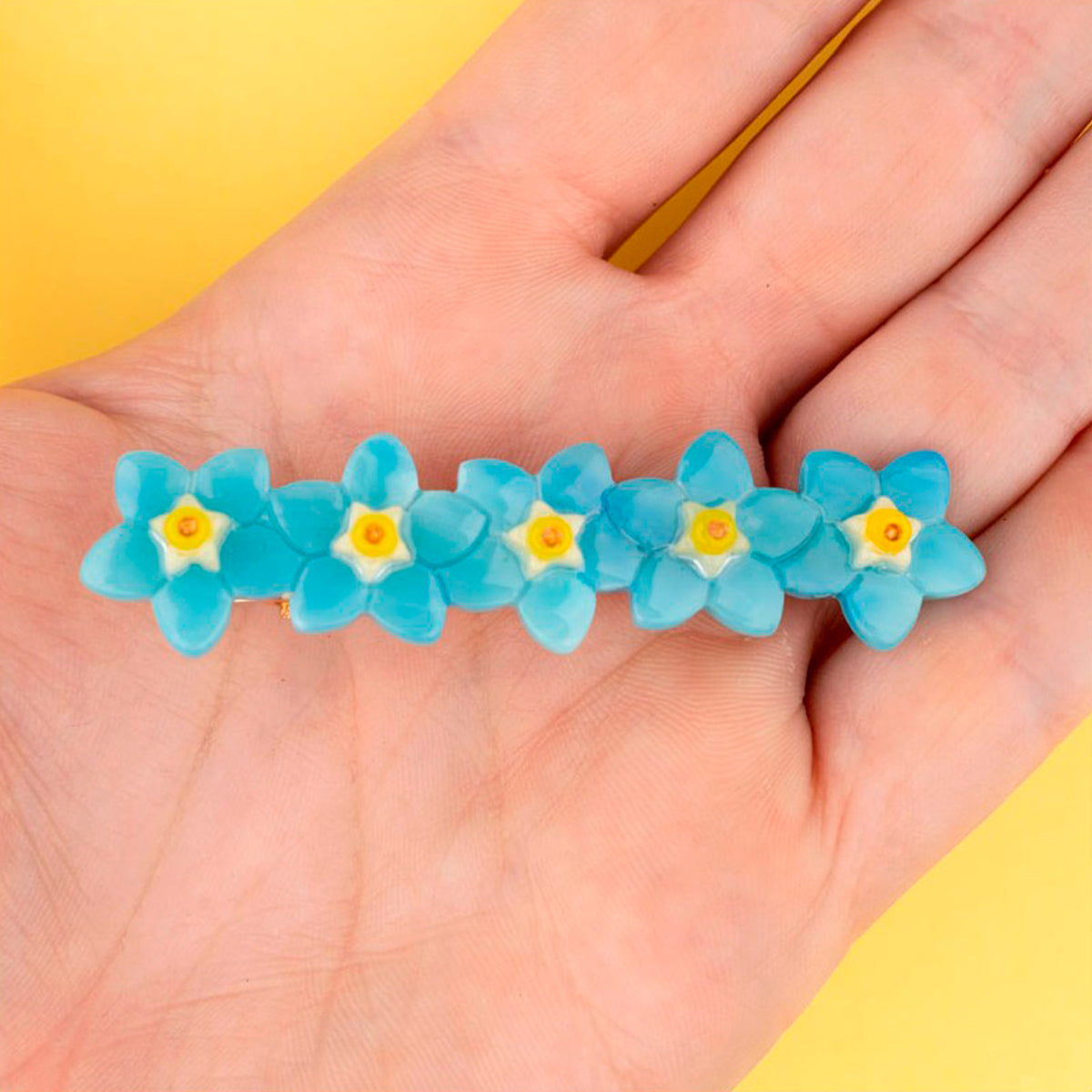 Pasador de pelo en forma de fila de flores nomeolvides azules con detalles en blanco y amarillo. Fabricado en acetato de celulosa reciclado. Vista sobre mano.
