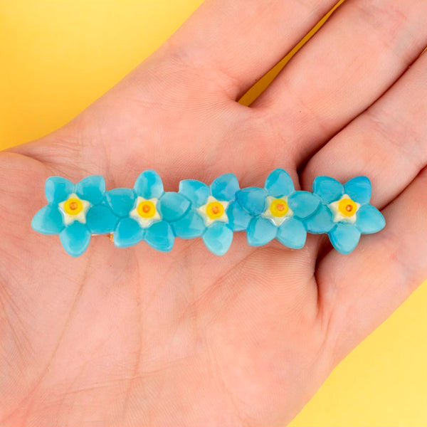 Pasador de pelo en forma de fila de flores nomeolvides azules con detalles en blanco y amarillo. Fabricado en acetato de celulosa reciclado. Vista sobre mano.