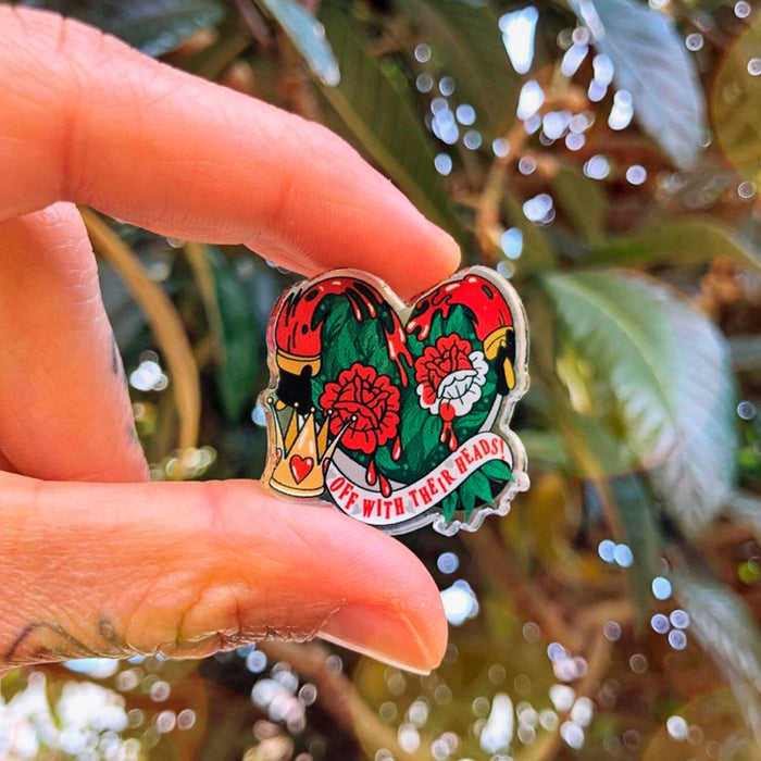 Pin acrílico con la frase "Off with their heads" ante un corazón hecho de hojas verdes, rosas pintadas de rojo, dos manos sujetando pinceles y una corona con un corazón.