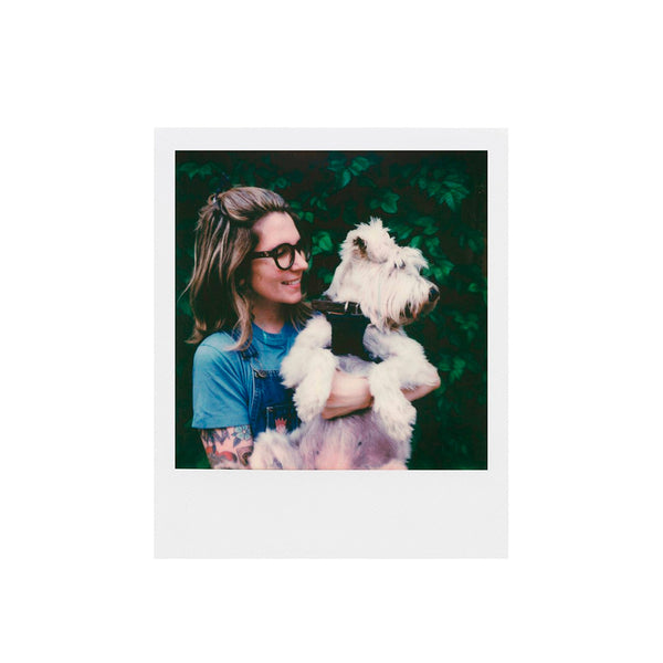 Fotografía polaroid a color i-type de una mujer sujetando en brazos a un perro ante un fondo de hojas verdes