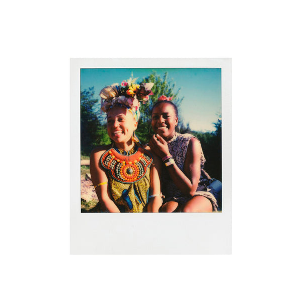 Fotografía polaroid a color i-type de dos mujeres negras sonriendo vestidas con un traje típico étnico en un fondo de naturaleza