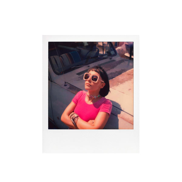 Fotografía polaroid sx-70 a color de mujer blanca con camiseta rosa y gafas de sol apoyada sobre un coche antiguo