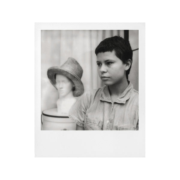 Fotografía polaroid i-Type en blanco  negro de una mujer racializada con pelo corto junto a un busto con sombrero