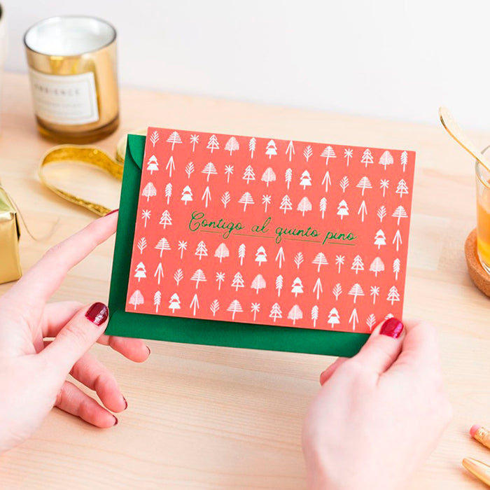 Fotografía de manos de mujer con uñas pintadas de rojo sujetando una postal de color  rojo con estampado de árboles geométricos y el texto "contigo al quinto pino" y sobre de color verde.